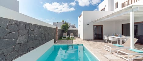 Un área de piscina moderna al aire libre con pared de piedra, sillas de descanso y zona de comedor con mesas y sillas.