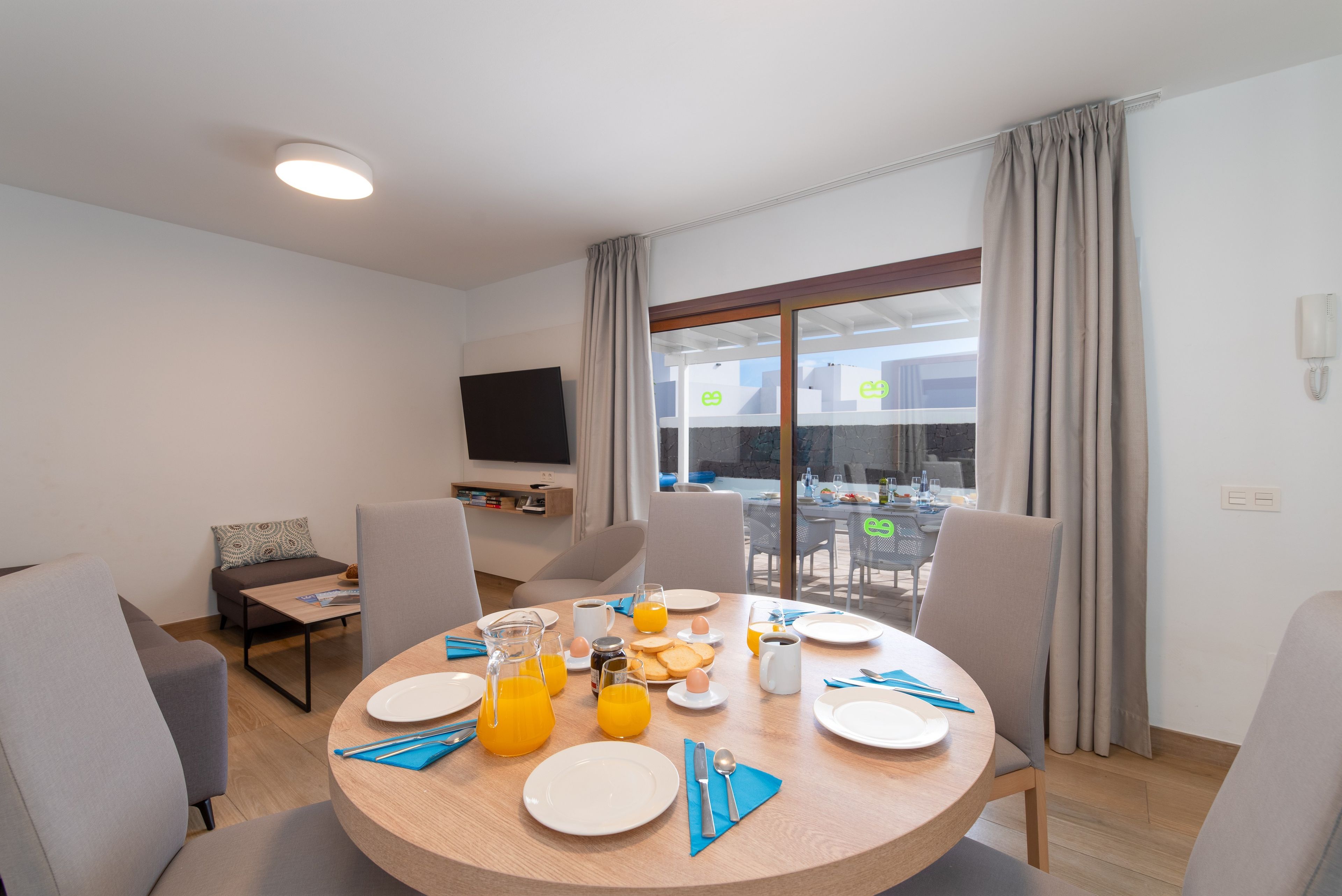 Un comedor con una mesa redonda dispuesta para el desayuno, un televisor en la pared y vistas al exterior a través de amplios ventanales.