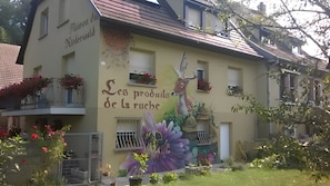 Un immeuble orné d’une fresque représentant un cerf et des fleurs, avec des balcons décorés de jardinières et une enseigne indiquant : « Produits de la ruche ».