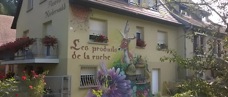 Un immeuble orné d’une fresque représentant un cerf et des fleurs, avec des balcons décorés de jardinières et une enseigne indiquant : « Produits de la ruche ».