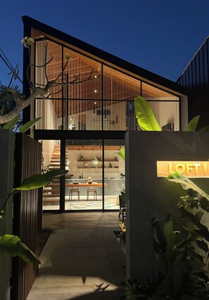 A modern two-story house with a glass facade, a visible kitchen area, and a dining space.