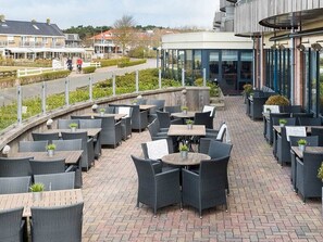 Een terras buiten met meerdere tafels en stoelen, een gebogen houten terras en een gebouw met grote ramen op de achtergrond.