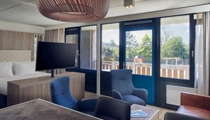 Een moderne hotelkamer met een groot bed, een flatscreen televisie, een zithoek met stoelen, en grote ramen met uitzicht op het buiten.