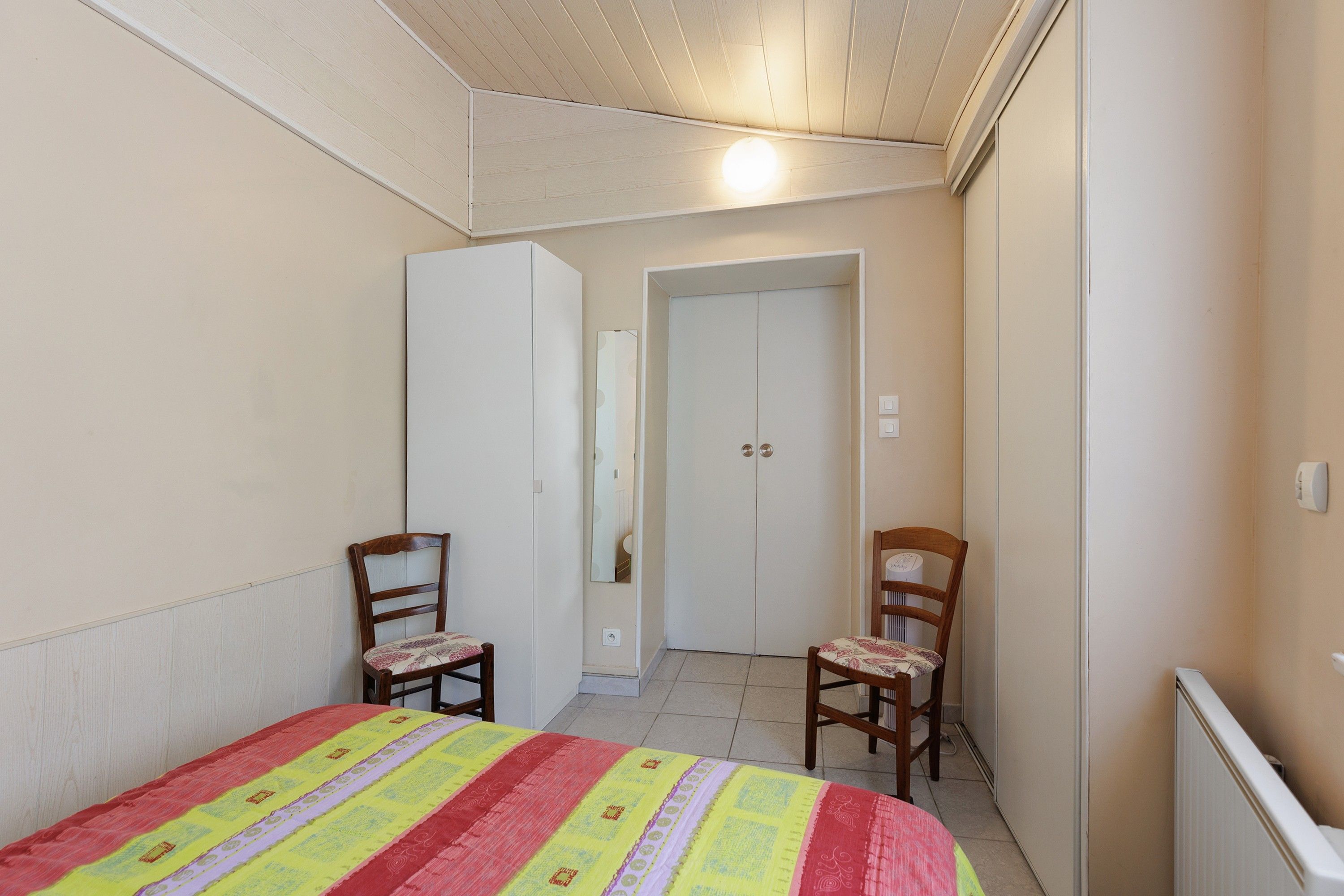 Une chambre à coucher comprenant un lit, deux chaises, une armoire et une porte donnant sur une autre pièce.