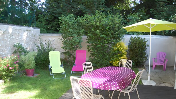 An outdoor patio with a table and chairs, surrounded by greenery and a white wall.