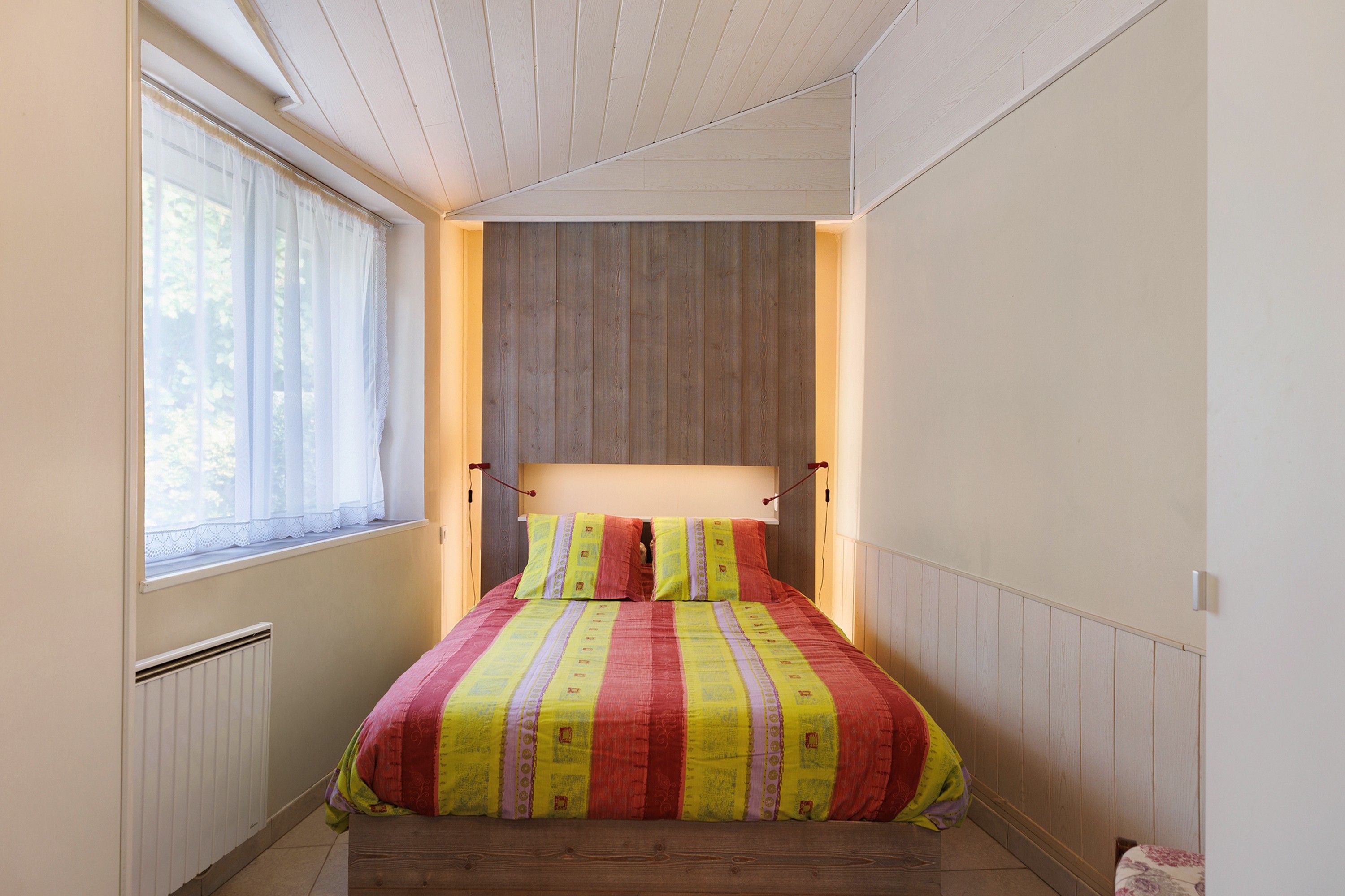 A bedroom with a wooden headboard, a window with sheer curtains, and a bed with striped bedding.