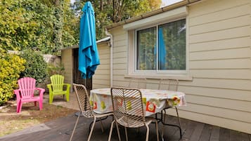 En hyggelig udendørs terrasse med et bord, stole, en blå parasol og et vindue med udsigt over en have.