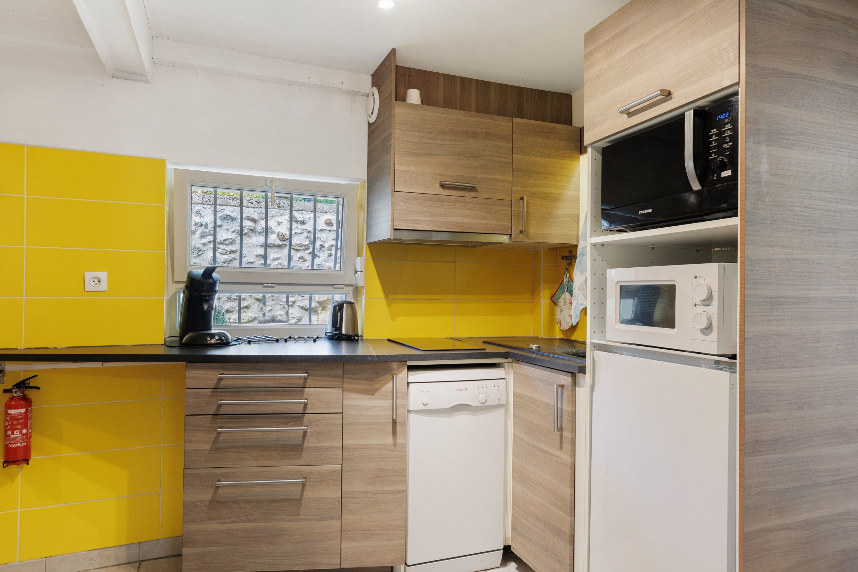 Una cocina moderna con armarios amarillos y blancos, un lavavajillas Bosch, un microondas Samsung y una ventana con vistas a un paisaje rocoso.