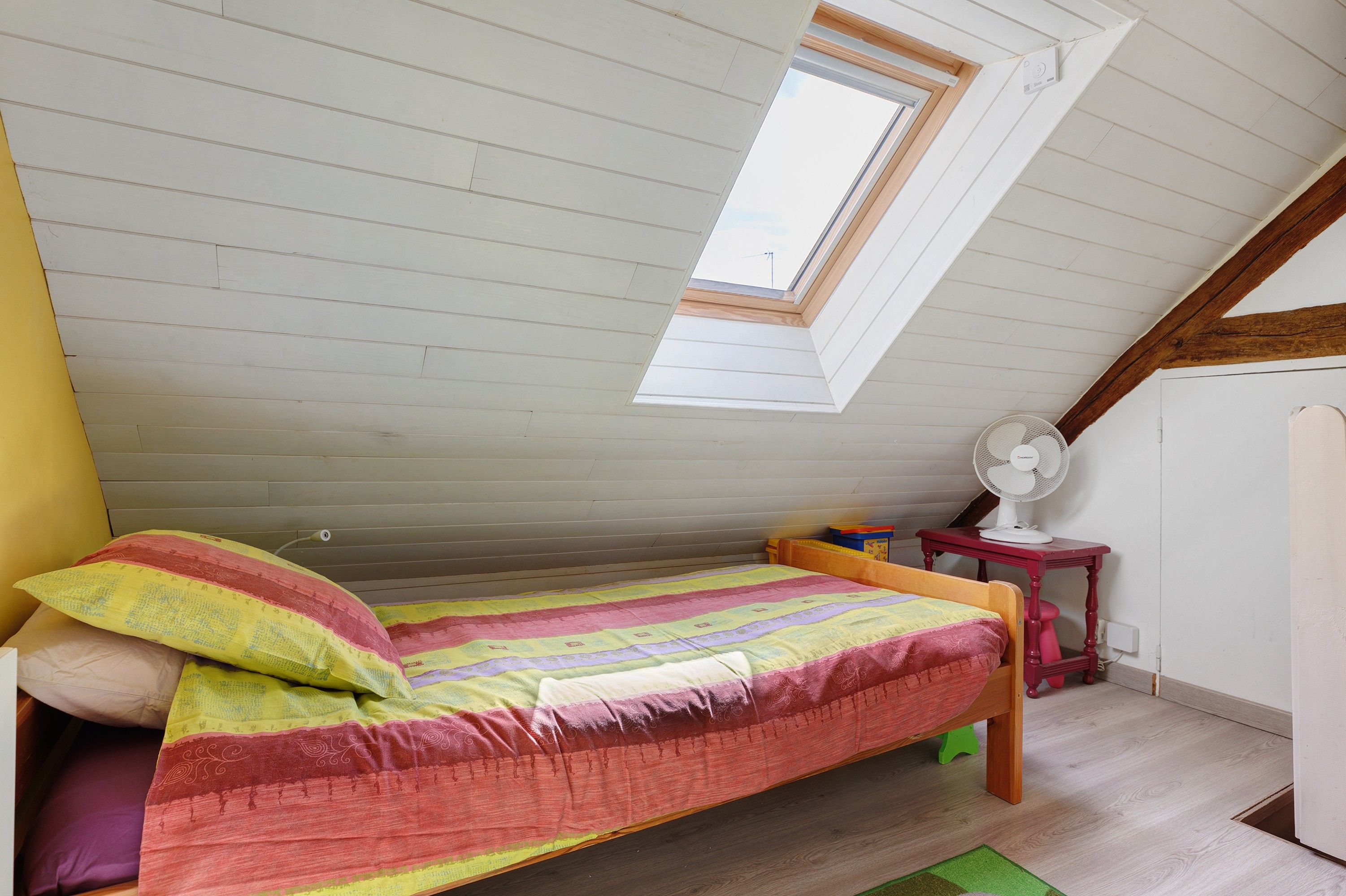 Ein kleiner Raum mit schrägem Dach, einem Einzelbett, einem Tisch, einem Ventilator und einem Fenster.
