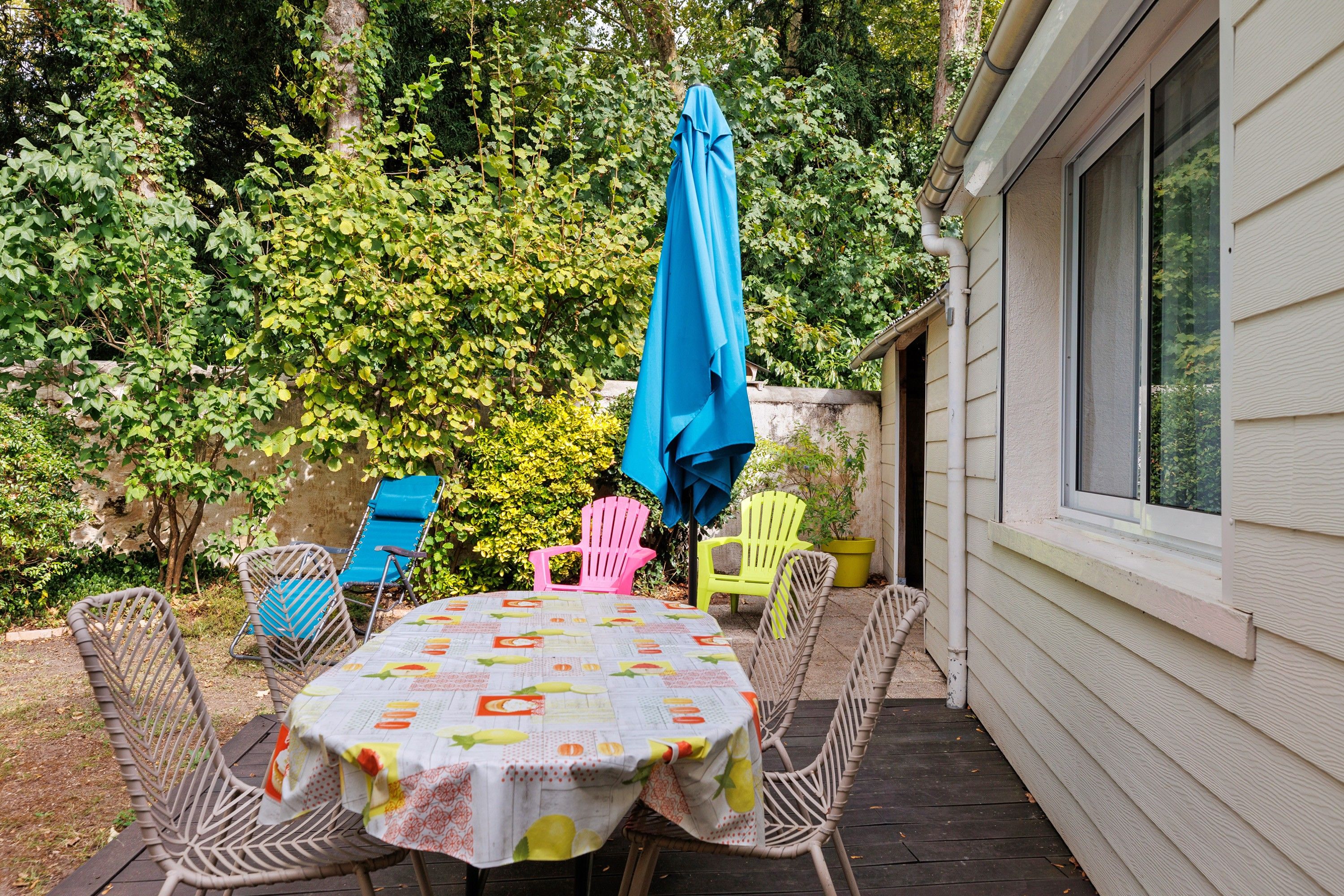 Un patio exterior con una mesa cubierta con un mantel de motivos frutales, rodeado de sillas de mimbre y un paraguas azul.