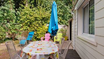 Un patio exterior con una mesa cubierta con un mantel de motivos frutales, rodeado de sillas de mimbre y un paraguas azul.
