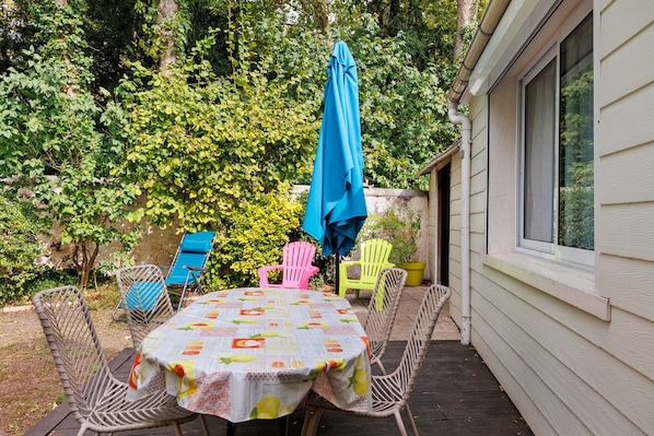 An outdoor patio with a table covered by a tablecloth with fruit patterns, surrounded by wicker chairs and a blue umbrella.