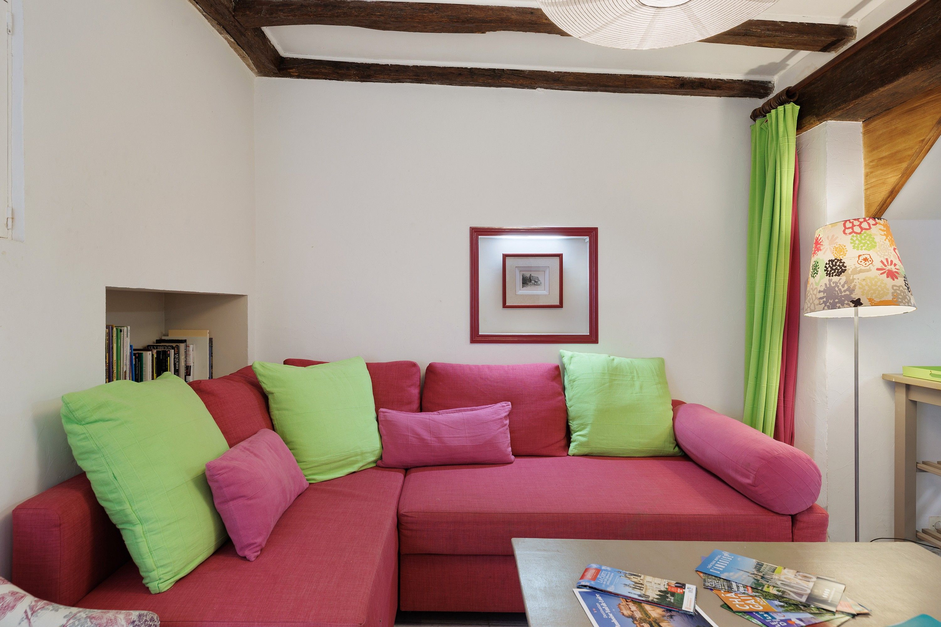 Una sala de estar con un sofá rojo, cojines verdes y rosados, un cuadro en la pared y una mesa con revistas.