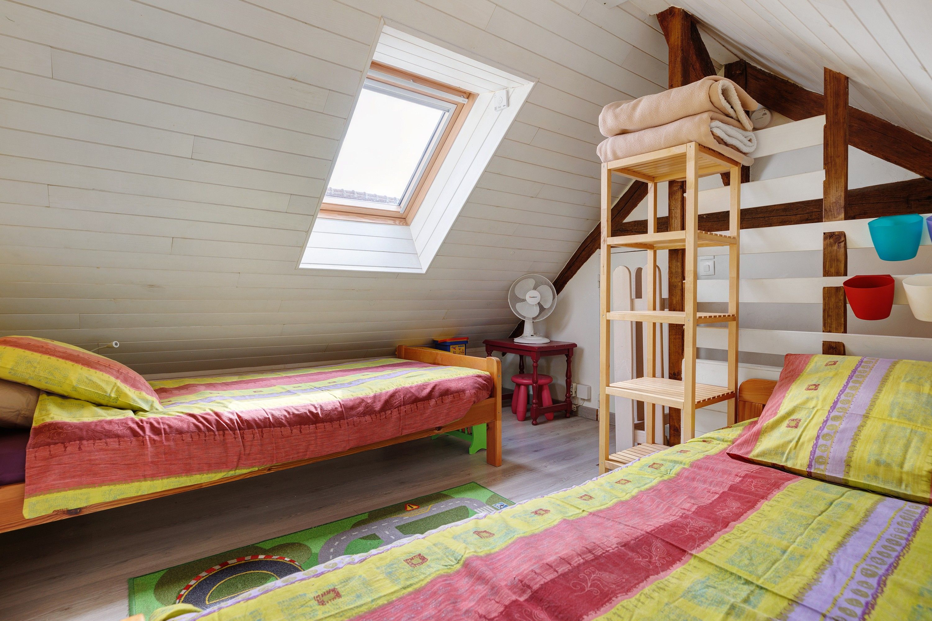 Een gezellig slaapkamer onder het dak met twee houten bedden, een dakraam, een ventilator en een stapelbed met opbergruimte.