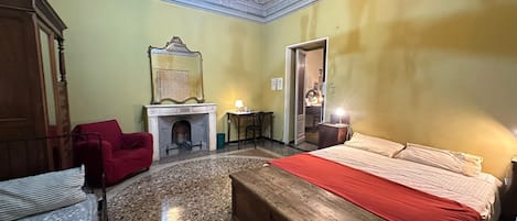 Une pièce comprenant un lit, un fauteuil rouge, une commode en bois, une cheminée et un lustre.