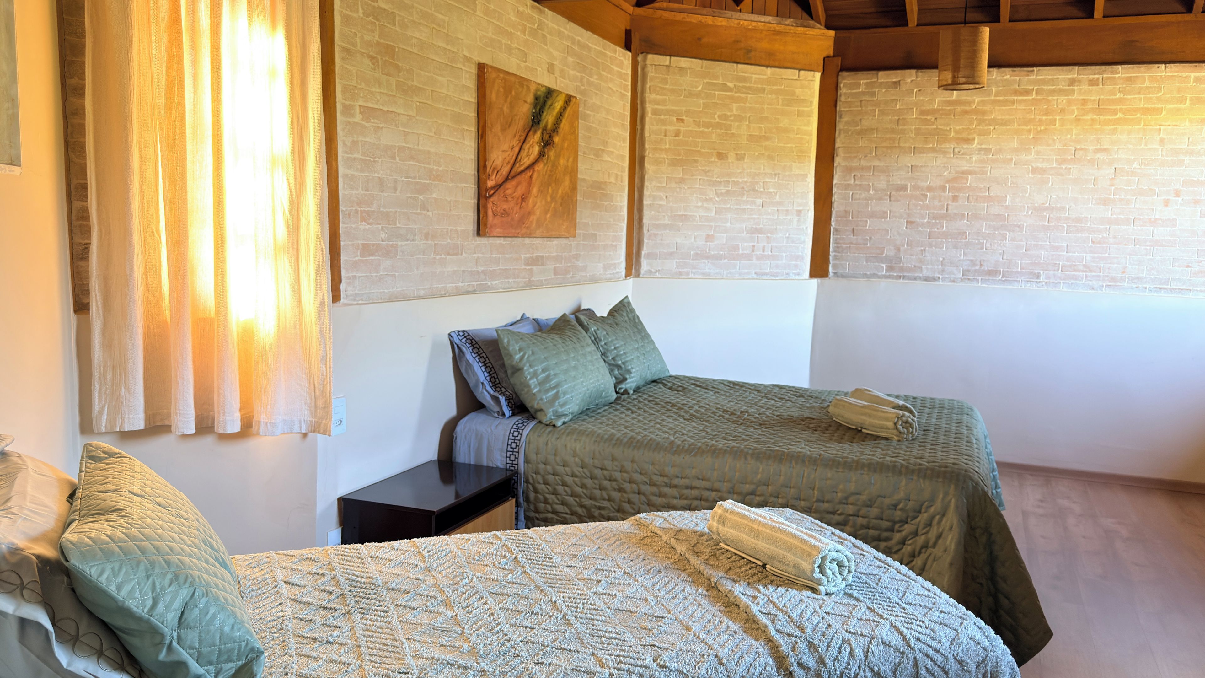 A room with two beds, a nightstand, a painting on the wall, and a window with curtains.