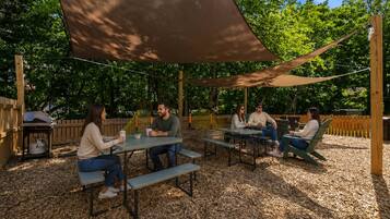 An outdoor seating area with tables and benches, surrounded by trees and a wooden fence.