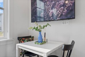 Une pièce avec une table blanche, un vase bleu rempli de fleurs, une chaise noire et un téléviseur fixé au mur.