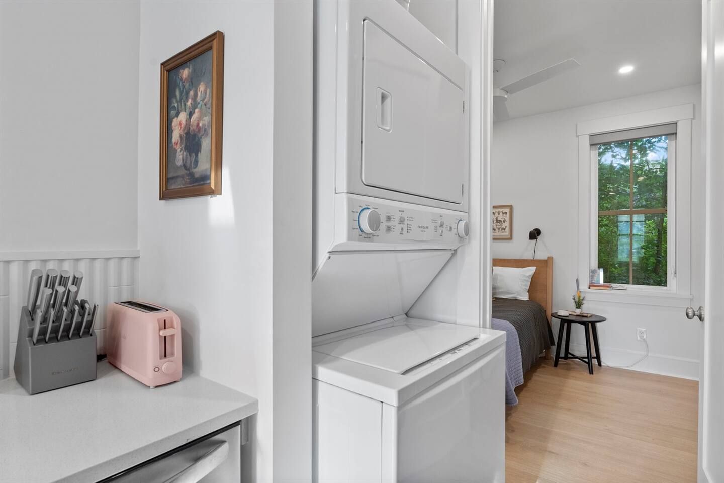 A compact room with a bed, a small table, a window with a view of trees, and a washing machine.