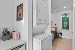 Quarto compacto com cama, uma mesinha, janela com vista para árvores e máquina de lavar.