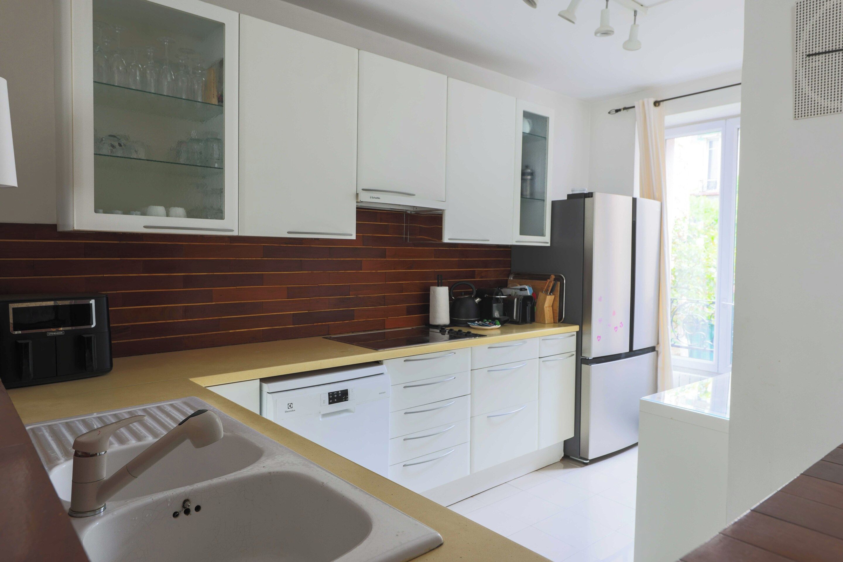 A modern kitchen with white cabinets, a stainless steel refrigerator, a dishwasher, and a window providing natural light.
