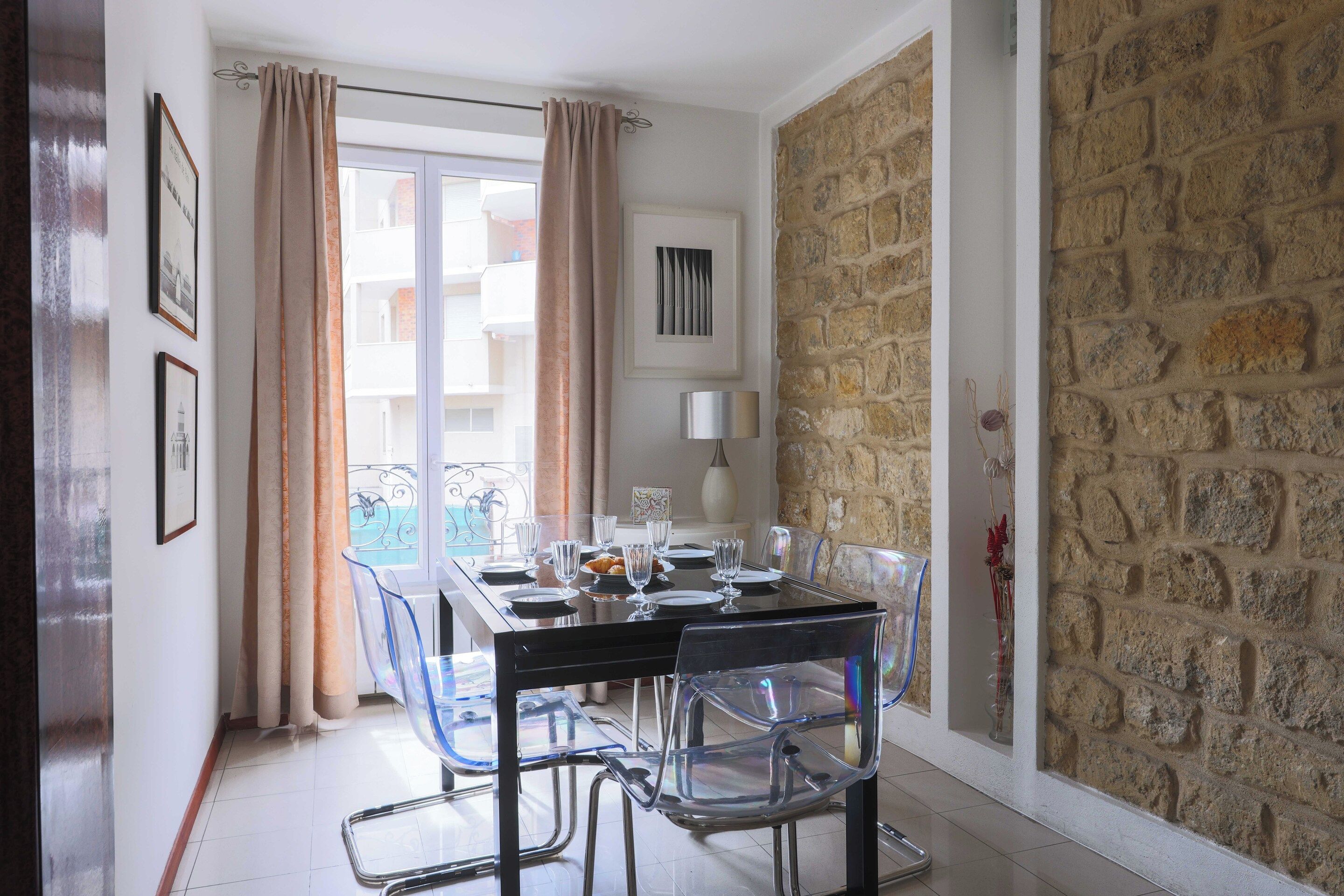 A dining area with a table set for four, clear chairs, a stone wall, and a balcony with a view.
