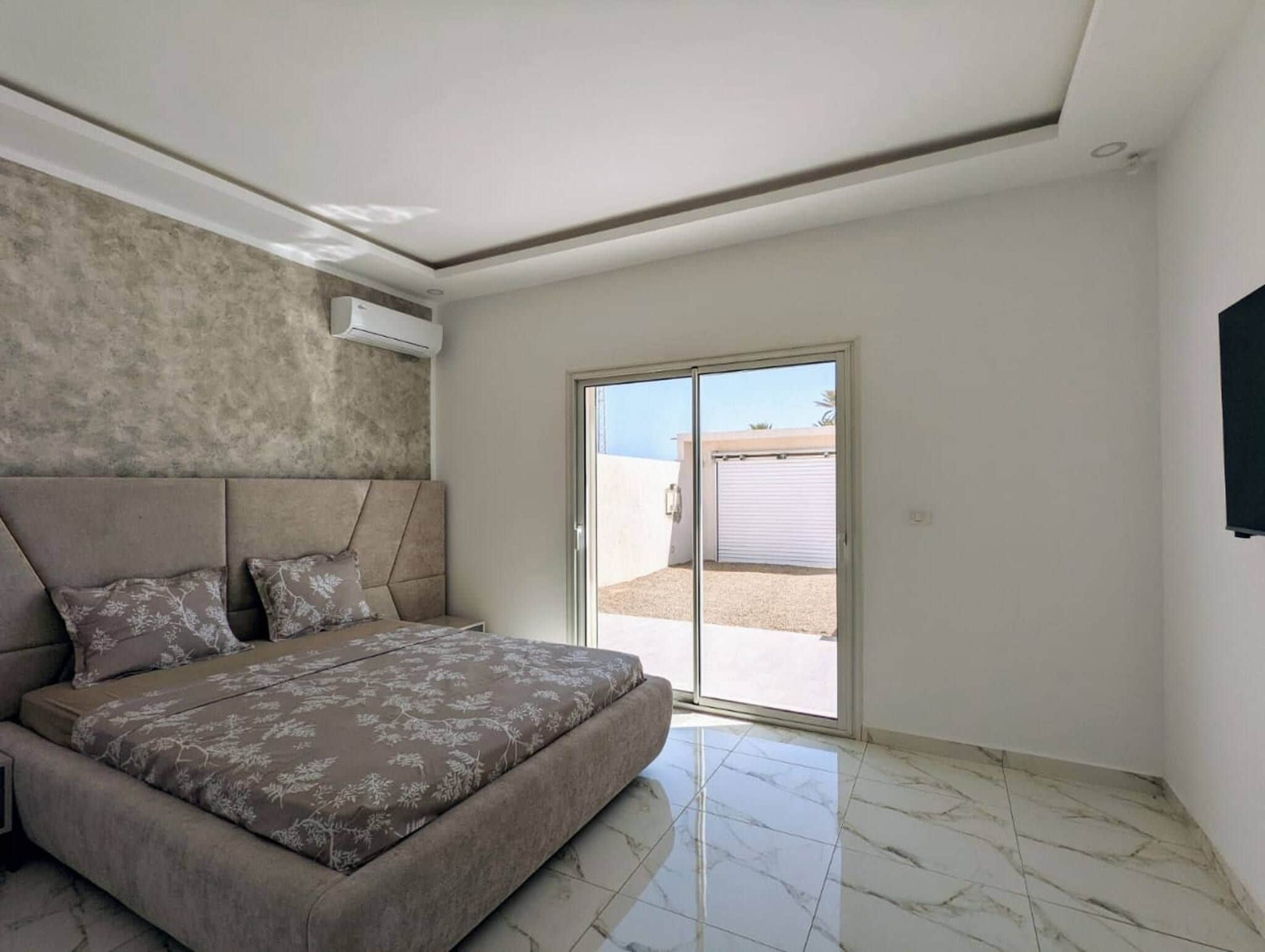 Une chambre dotée d’un grand lit, d’une tête de lit intégrée, d’une porte-fenêtre coulissante et d’un appareil de climatisation fixé au plafond.