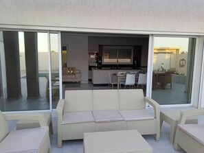 A covered outdoor seating area with a sofa and chairs, a dining table with chairs, and a kitchen area visible through glass doors. - Escape to Villa Yasmina (Aghir)