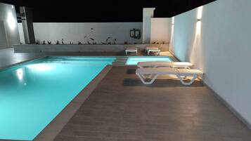 Una piscina exterior con solárium de baldosas y sillas reclinables blancas.