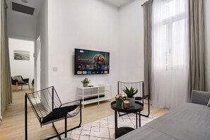 A modern hotel room with a bed, a TV displaying 'The Wheel of Time' series, a small table with a plant, two chairs, and a white TV stand. - The Monti – Stylish 1‑Bedroom Apartment in Central Tel Aviv (Tel Aviv-Yafo)