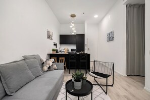 A modern living room with a grey sofa, a black coffee table with plants, and a black chair. A kitchen with black cabinets and a white countertop is visible in the background. - The Monti – Stylish 1‑Bedroom Apartment in Central Tel Aviv (Tel Aviv-Yafo)