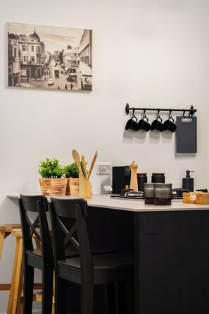 A kitchen with a black countertop, wooden chairs, a potted plant, and a vintage cityscape painting. - The Monti – Stylish 1‑Bedroom Apartment in Central Tel Aviv (Tel Aviv-Yafo)
