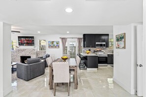 A modern living room with a fireplace, a dining area with a table and chairs, and a kitchen with dark cabinets and modern appliances. - West Palm Pool Home Sleeps 10 Game Room (West Palm Beach)