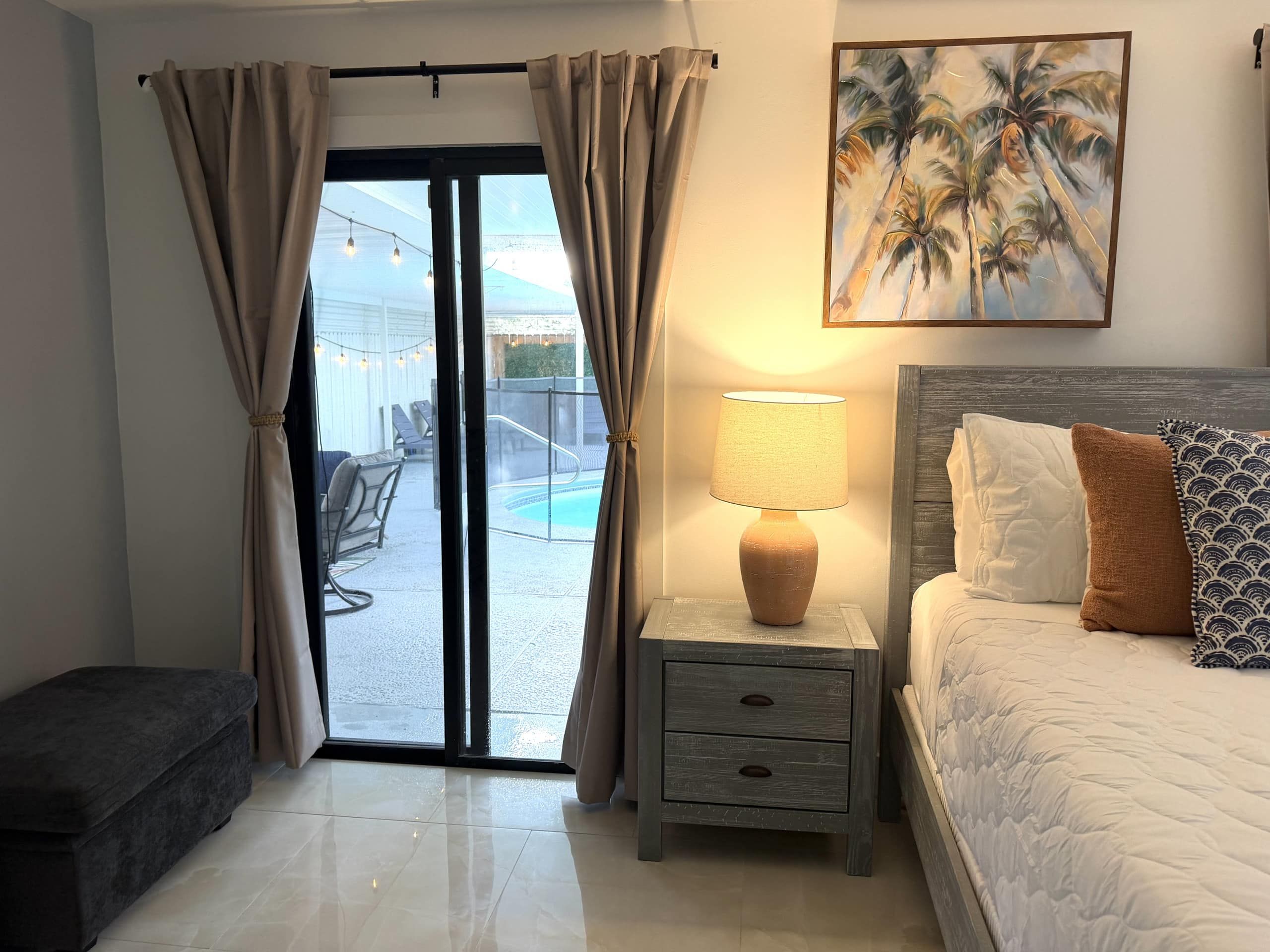 Habitación de hotel con cama, mesita de noche con lámpara, un cuadro de palmeras y vista a un área de piscina exterior.