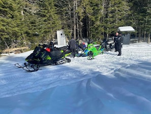 A snowy landscape with snowmobiles and a small building.