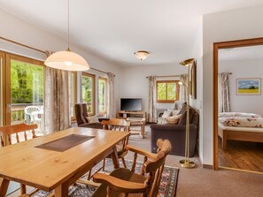 Ein großzügiger Wohnbereich mit Esstisch aus Holz, Stühlen, einer Couch, einem Fernseher und großen Fenstern mit Blick auf Grünflächen.