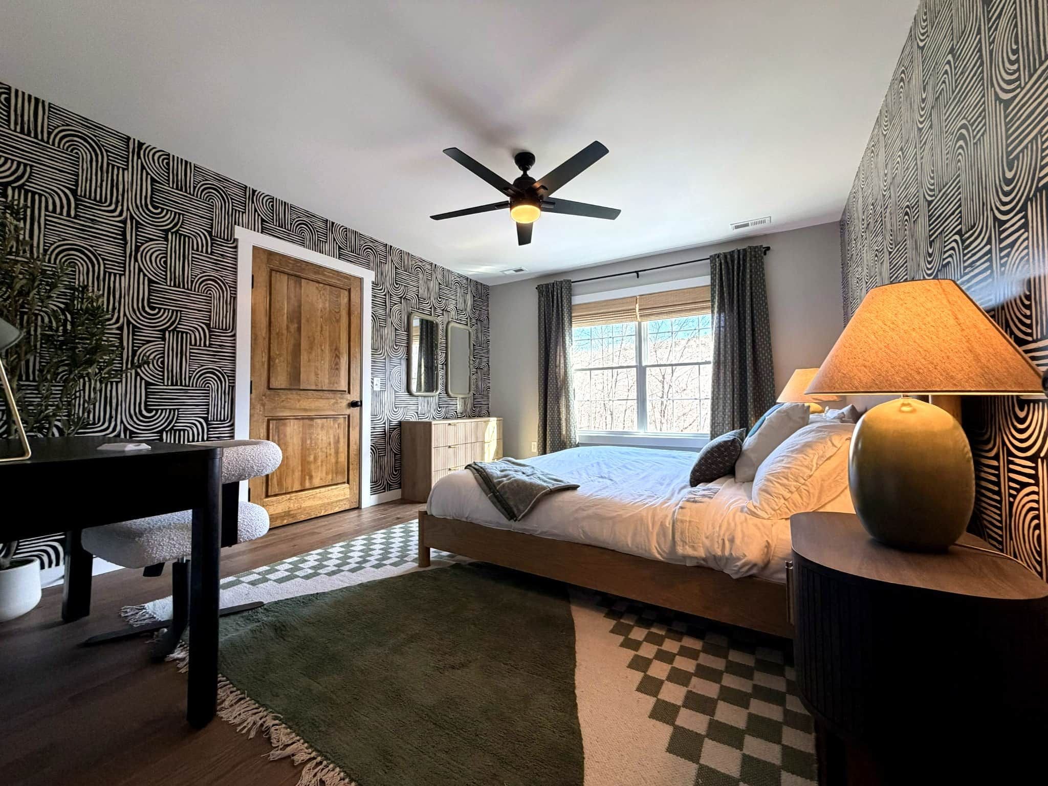 Une chambre à coucher comprenant un lit, un bureau, une chaise, une lampe, un ventilateur de plafond et une fenêtre avec des rideaux.