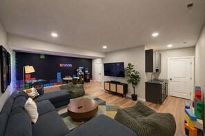 Una sala de estar moderna con sofá, reposapiés, televisor, consola de videojuegos y una pequeña zona de cocina.