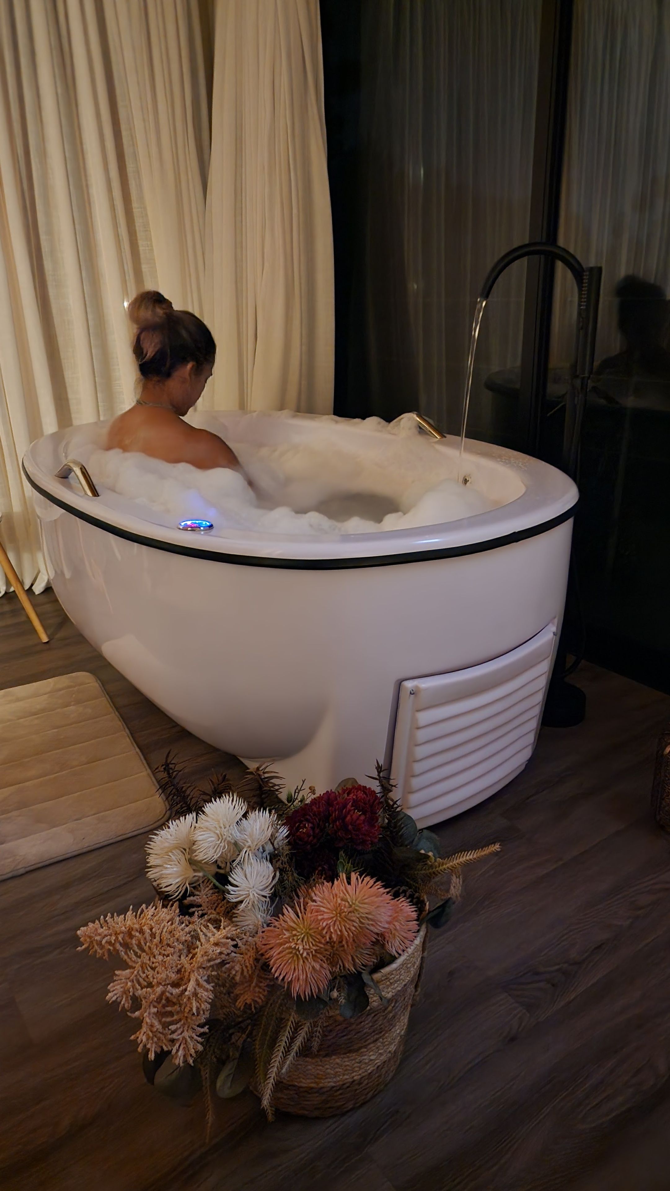 En person sitter i ett vitt bubbelbad med en kontrollpanel, omgiven av ett trägolv, en vit stol och en korg med blommor.