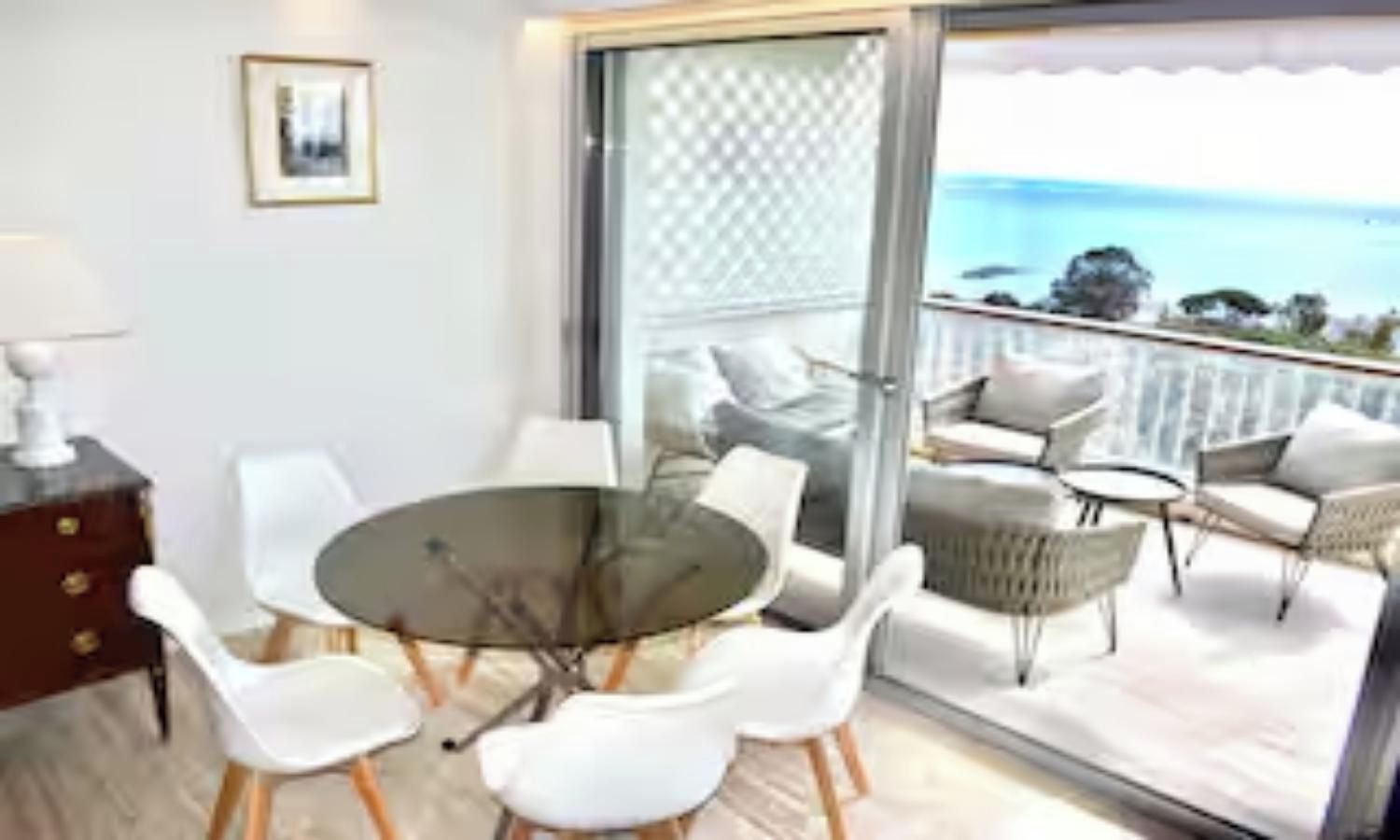 A dining area with a round table and chairs, a small side table, and a view of the sea through a glass door.