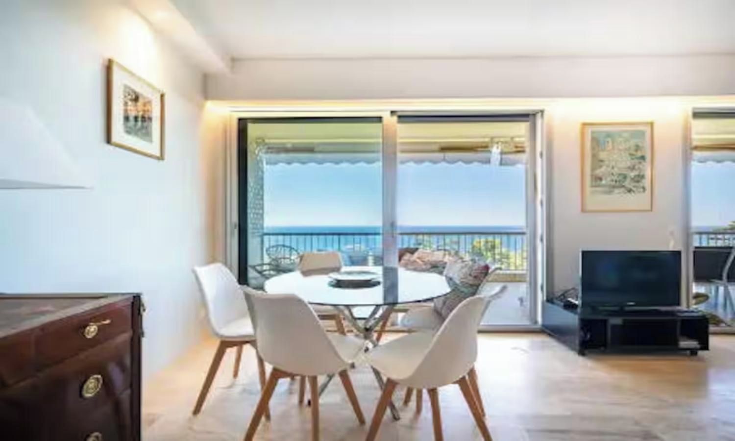 A dining area with a round table and chairs, a television, and a view of the sea through large sliding doors.