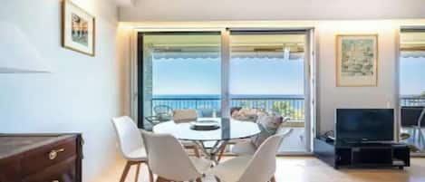 A dining area with a round table and chairs, a television, and a view of the sea through large sliding doors.