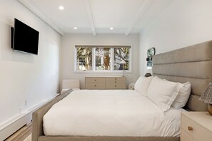 Una camera da letto con un letto, un comodino, una cassettiera, un televisore montato a parete e una finestra vista alberi.