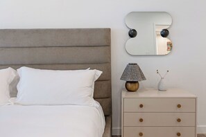 A bed with white bedding, a grey upholstered headboard, a bedside table with a lamp, and a wall-mounted mirror. - Villa Cortina 135 | 2BR Vail Village Retreat with Pool (Vail)