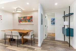 Un'area pranzo moderna con un tavolo e sedie in legno, un quadro appeso al muro e un corridoio che conduce a un'altra stanza.