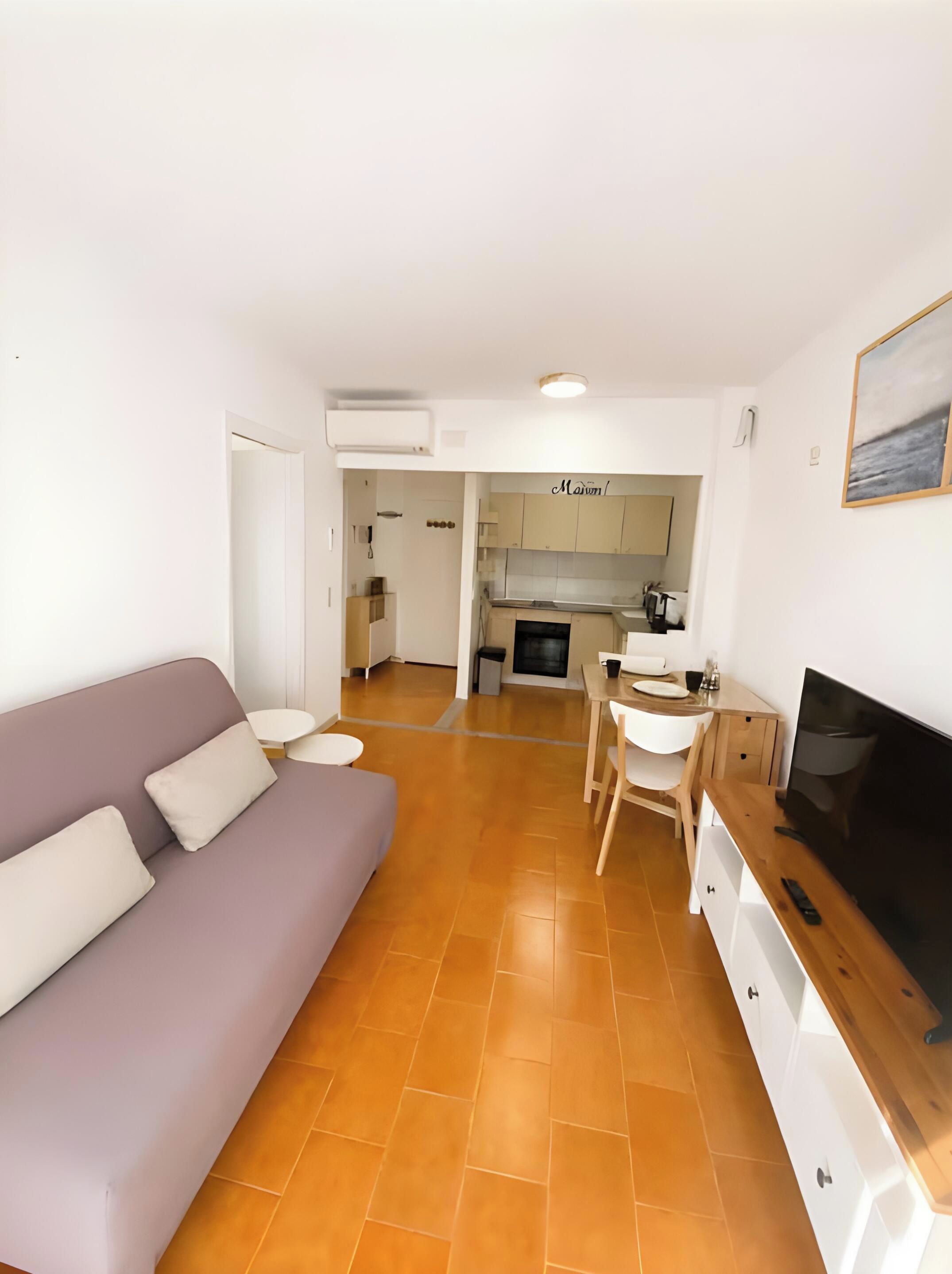 Una sala moderna con sofá, un comedor con mesa y sillas, una cocina al fondo y un televisor de pantalla plana sobre un soporte de madera.