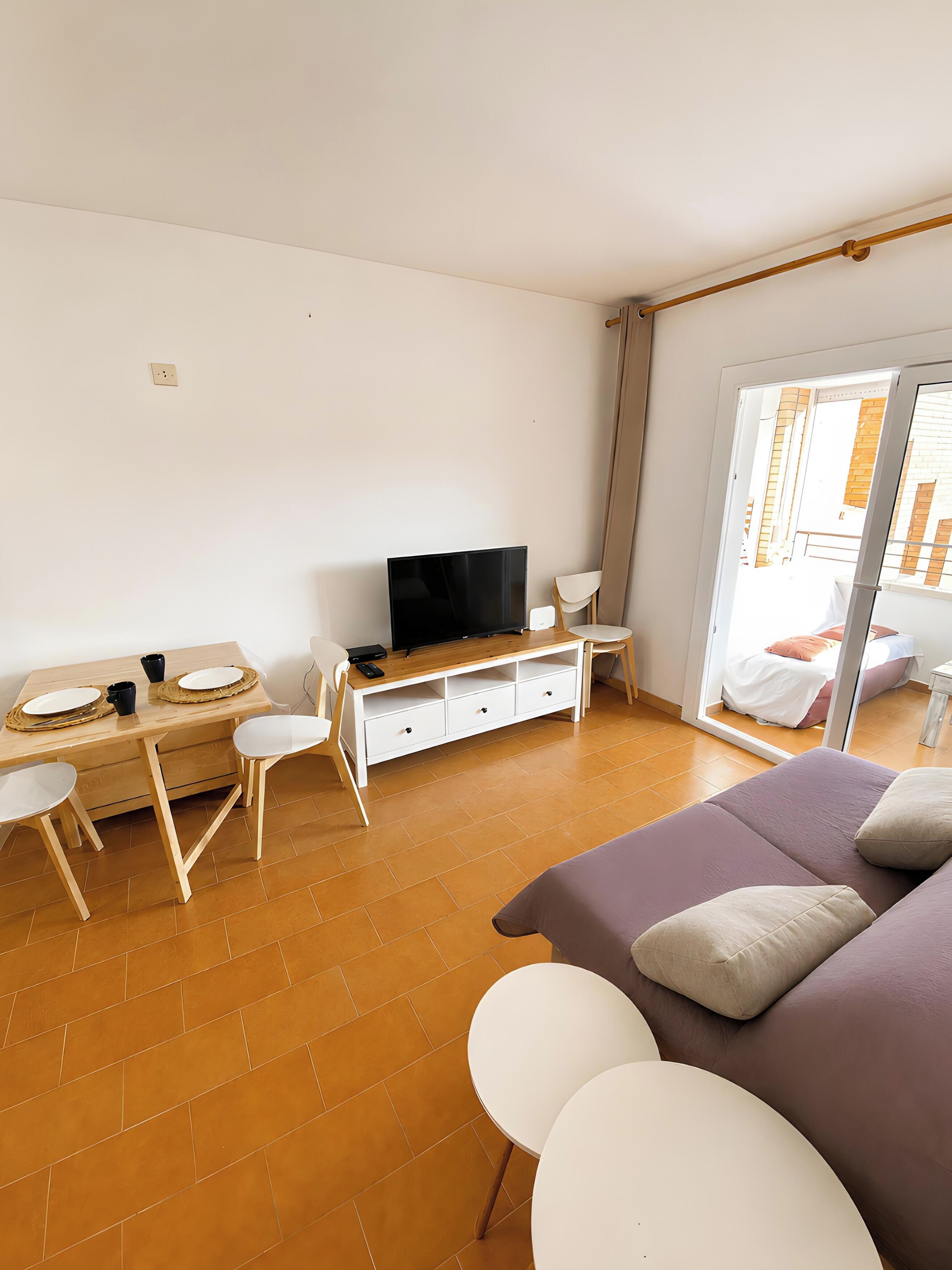 A room with a bed, a TV, a desk with chairs, and a sliding glass door to a balcony.