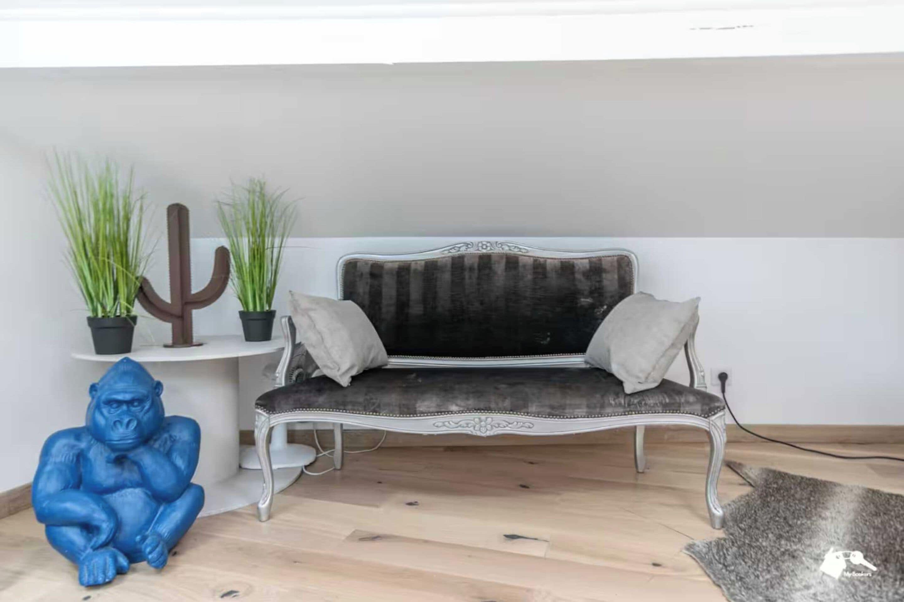 A room with a dark grey upholstered bench, two white cushions, a cactus, and a blue gorilla statue.