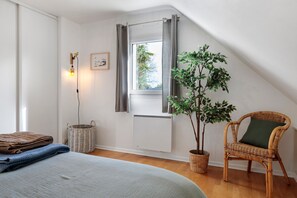 Un dormitorio con cama, una silla de mimbre, una planta en maceta, una ventana con cortinas y un aplique de pared.