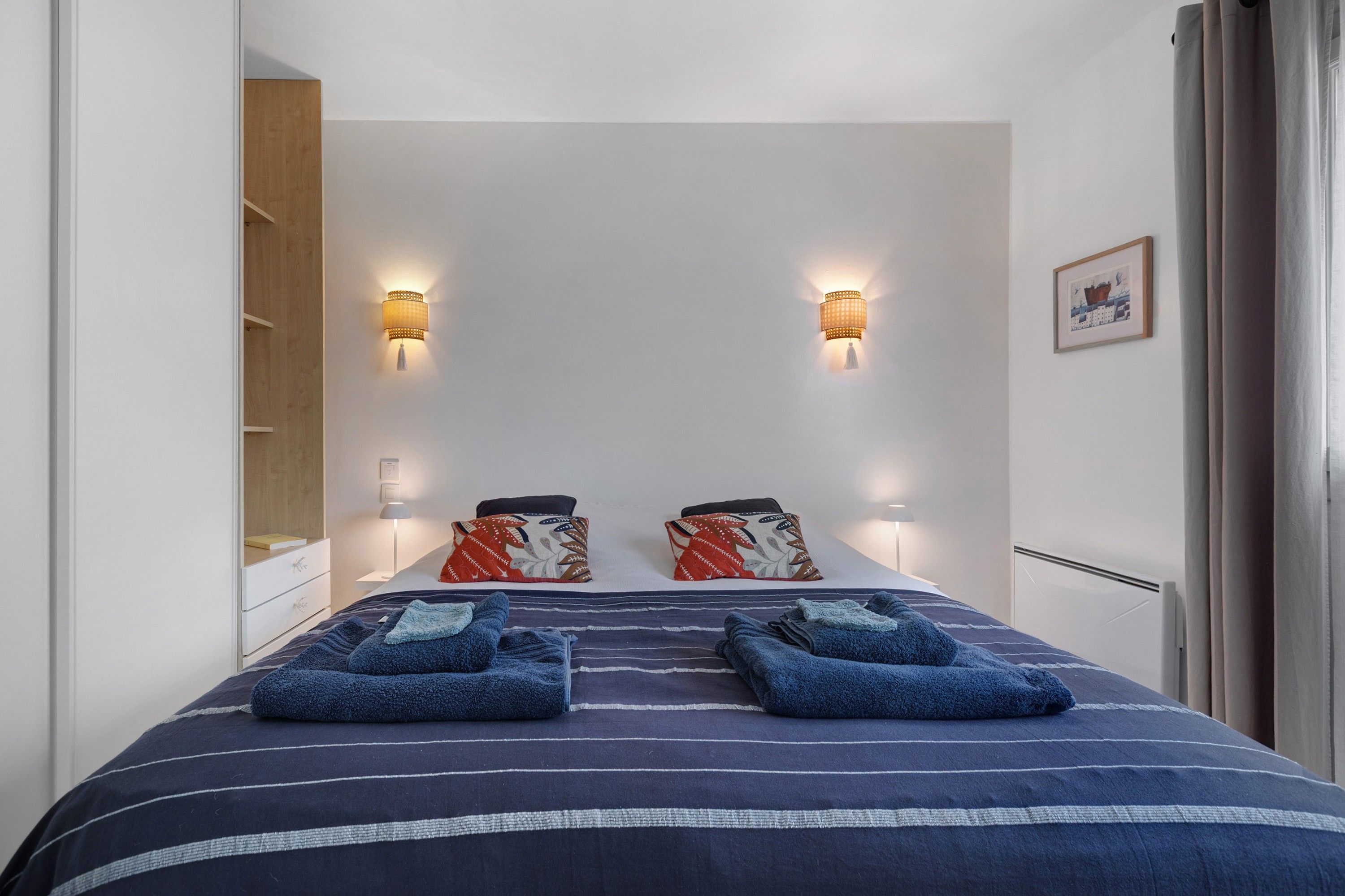 A hotel room with a bed, two wall-mounted lamps, a framed picture, and a window with curtains.